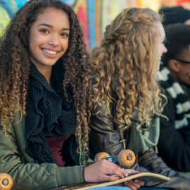 A multi-ethnic group of teenagers are sitting against a wall covered in graffiti. A female of African descent looks away from her friends and smiles as she holds a skateboard.